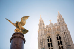 A golden eagle statue with outstretched wings is mounted on a tall, dark pedestal in the foreground. In the background, there is a large Gothic-style building with tall spires against a clear blue sky.