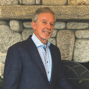 A man with gray hair and a beard, dressed in a dark suit jacket and light blue shirt, stands in front of a stone fireplace, smiling slightly at the camera in anticipation of Fall 2025.