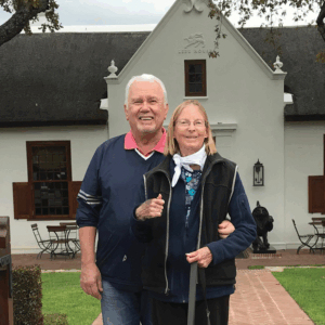 An older man and woman stand arm in arm, smiling in front of a white building with wooden doors and windows. Outdoor chairs and tables rest on the brick patio, surrounded by grass, capturing a warm Fall 2025 afternoon.