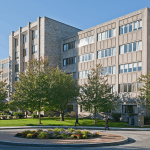 A large, multi-story academic building with many windows, trees, and a circular flower bed in front. Two people walk on the path near the building under a clear blue sky, hinting at the vibrant campus life of Fall 2025.